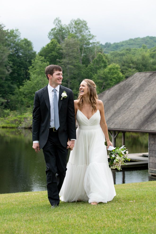 A couple in wedding attire walking and laughing together on grass near a lake and wooden boathouse on their special day.