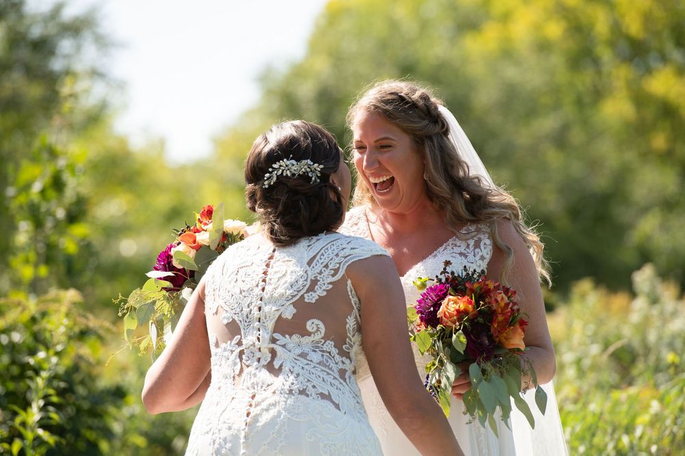 Two people in white lace dresses embrace joyfully surrounded by sunny greenery during a wedding celebration.