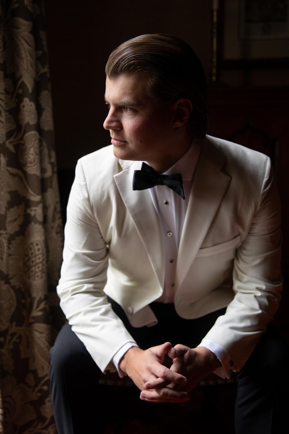 Groom in white tuxedo jacket sitting thoughtfully in dramatic window lighting.