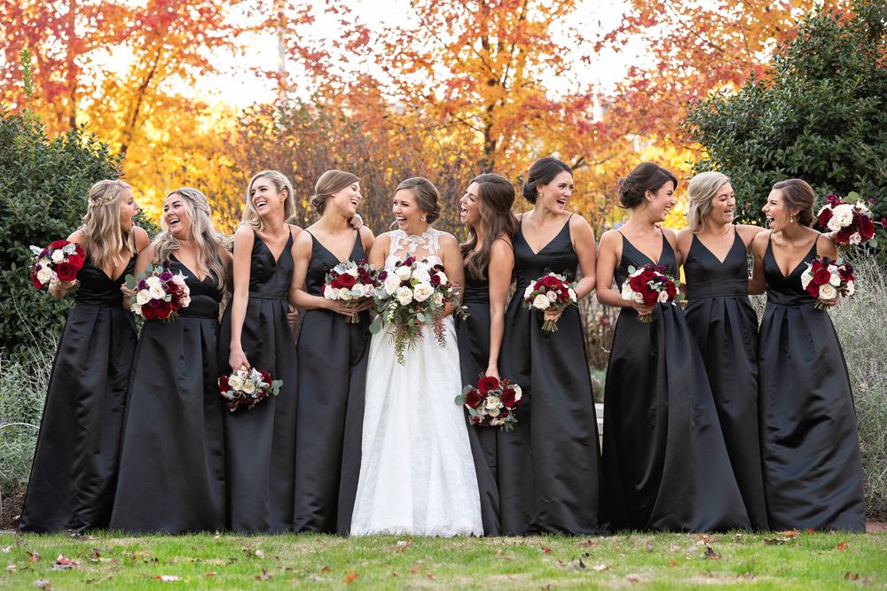 Wedding party wearing black bridesmaid dresses stands together outdoors against autumn foliage at sunset.