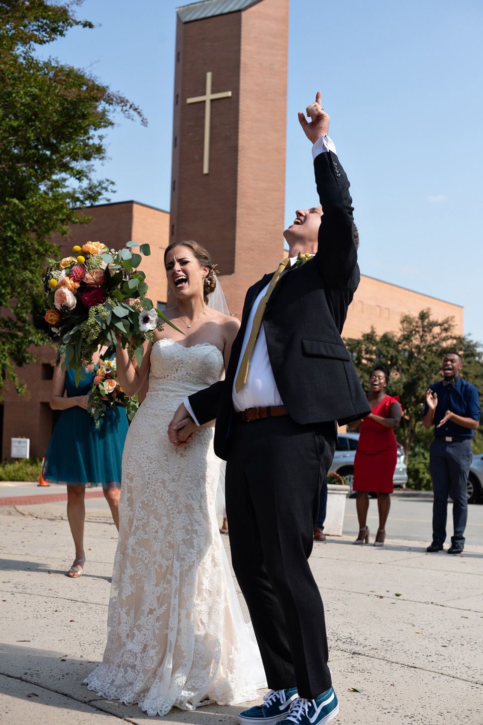 A joyful wedding celebration outside a church with a tall cross-adorned bell tower in the background.