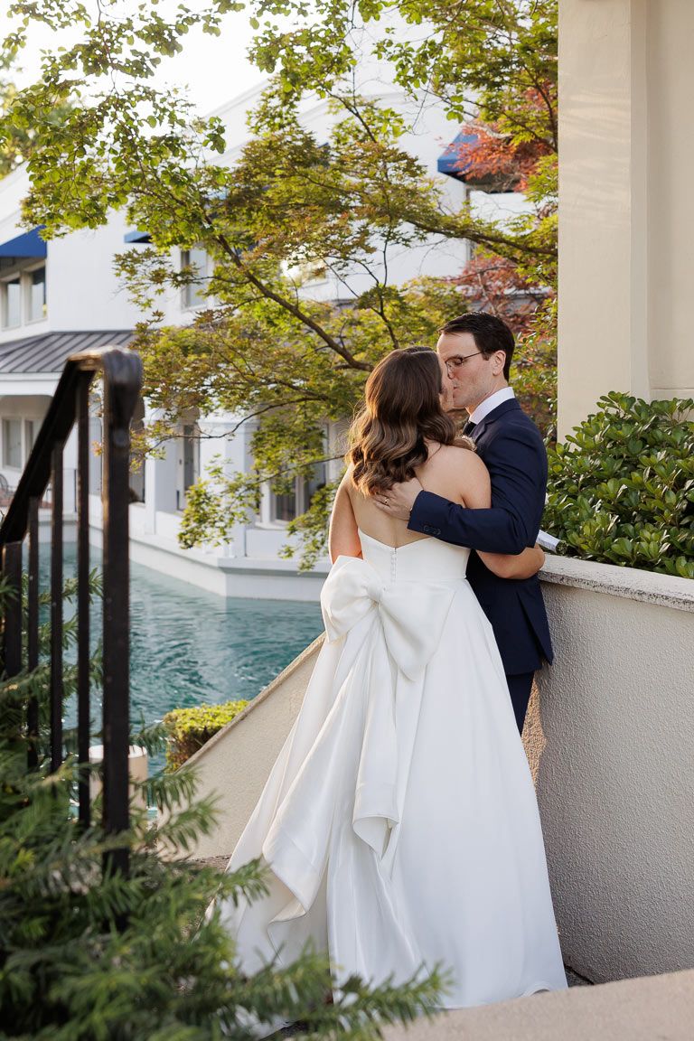 Elegant bride and groom share their first look moment under a columned portico with lush greenery.