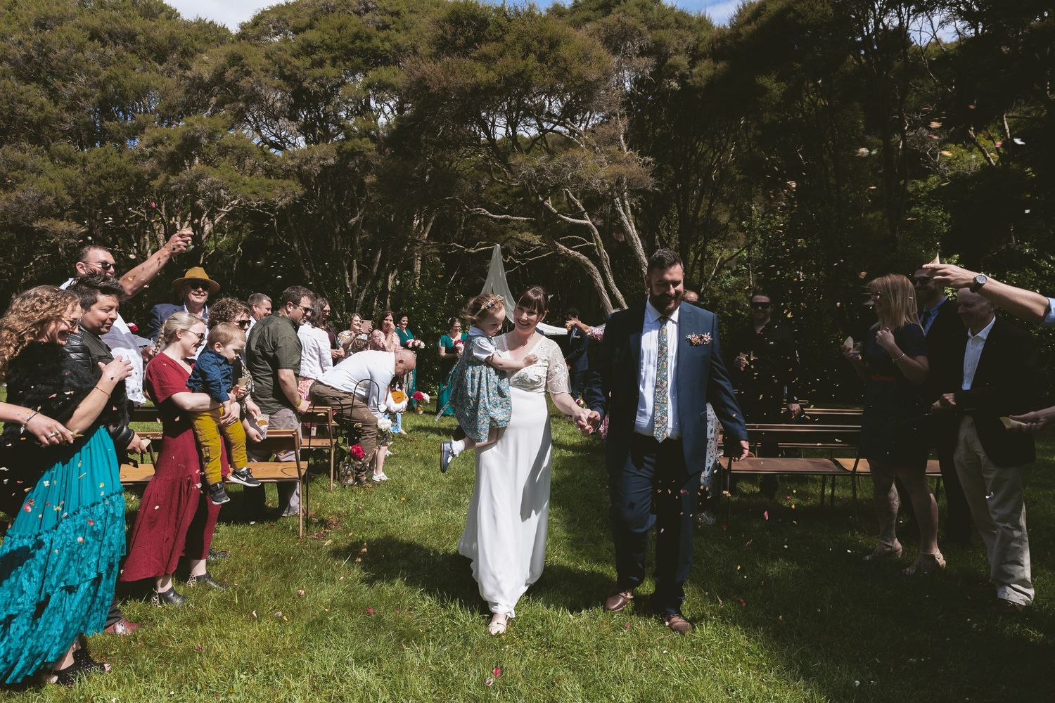 Wedding guests celebrate with raised hands as couple walks down outdoor aisle lined with wooden chairs.
