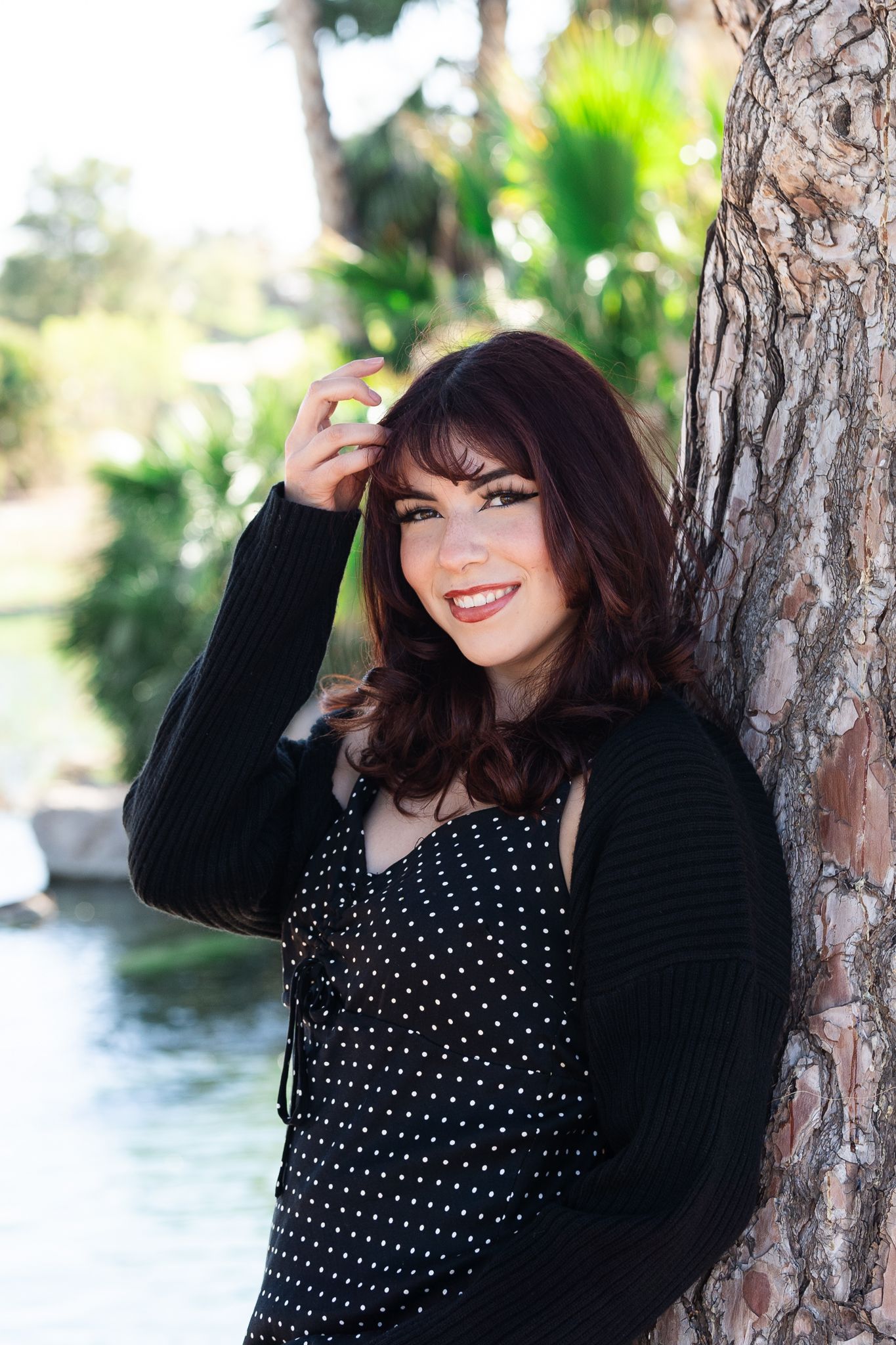 Casual portrait leaning against a tree trunk wearing a spotted dress and black cardigan.