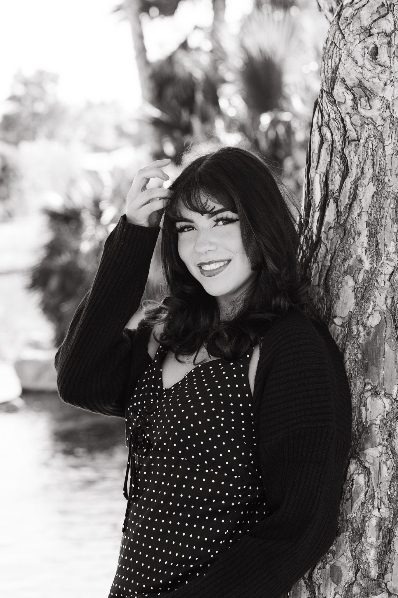 A black and white portrait of someone in a polka dot dress and cardigan leaning against a tree.