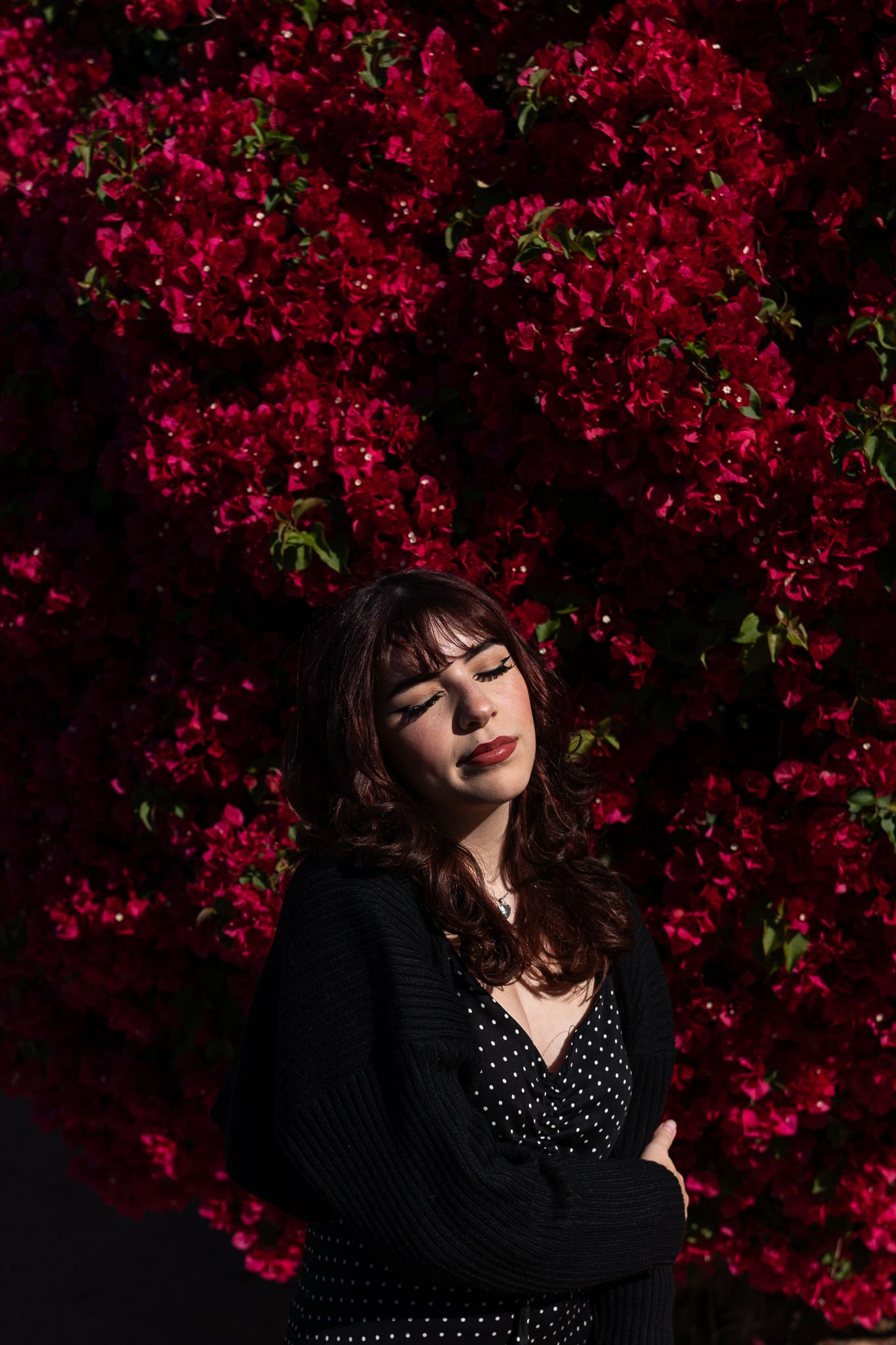 Moody portrait against dark red bougainvillea in black polka dot dress.