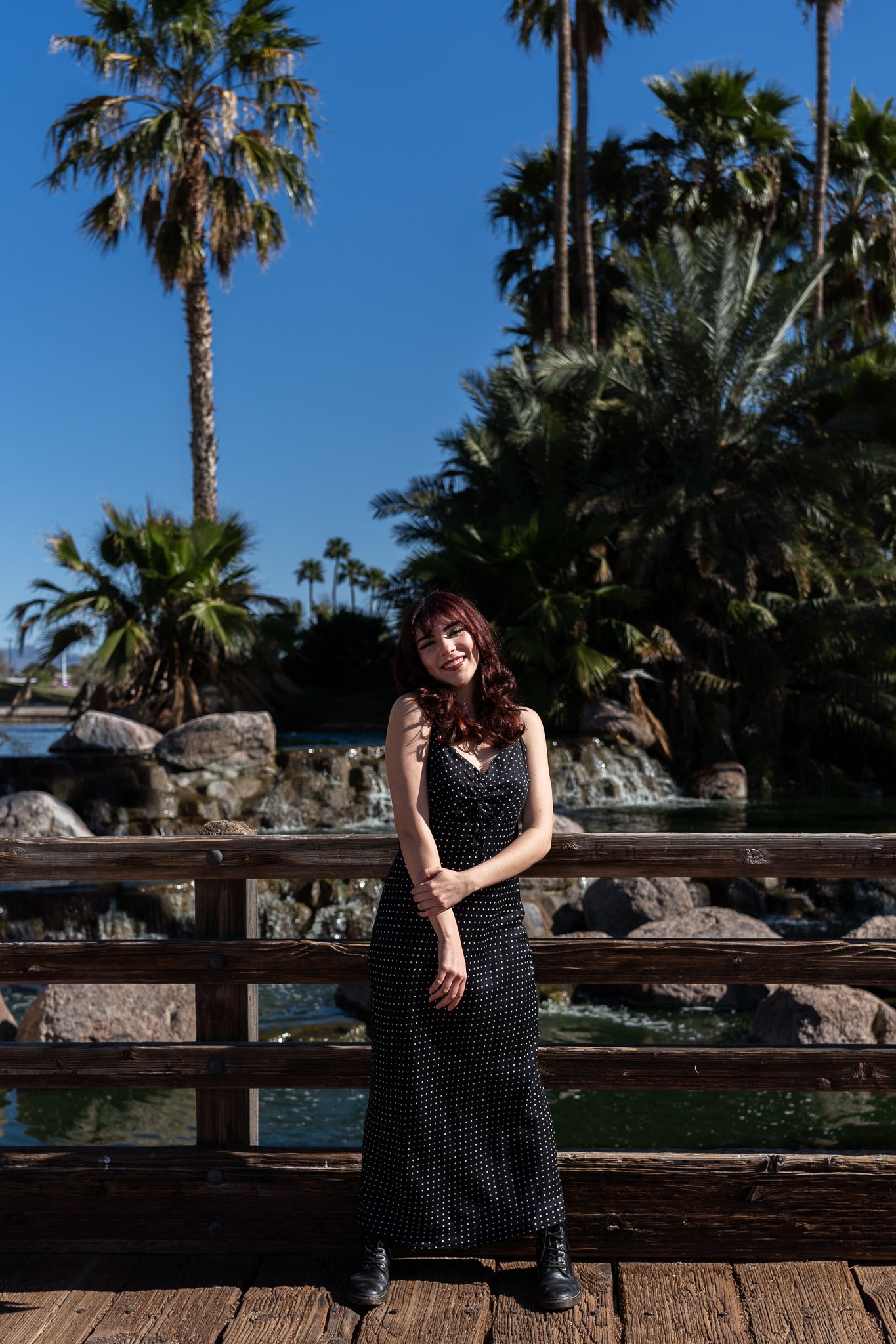Full length portrait on wooden deck with palm trees and water feature in background.