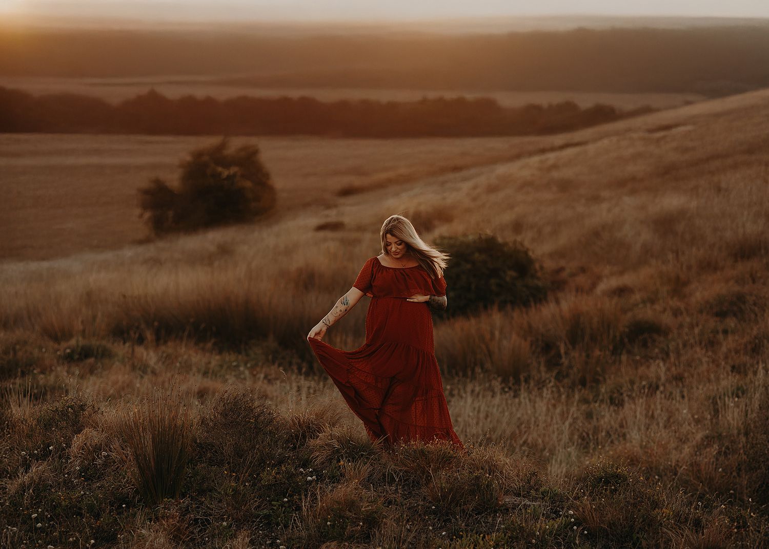 Woman in flowing red dress twirling in golden sunset light on grassy hillside.