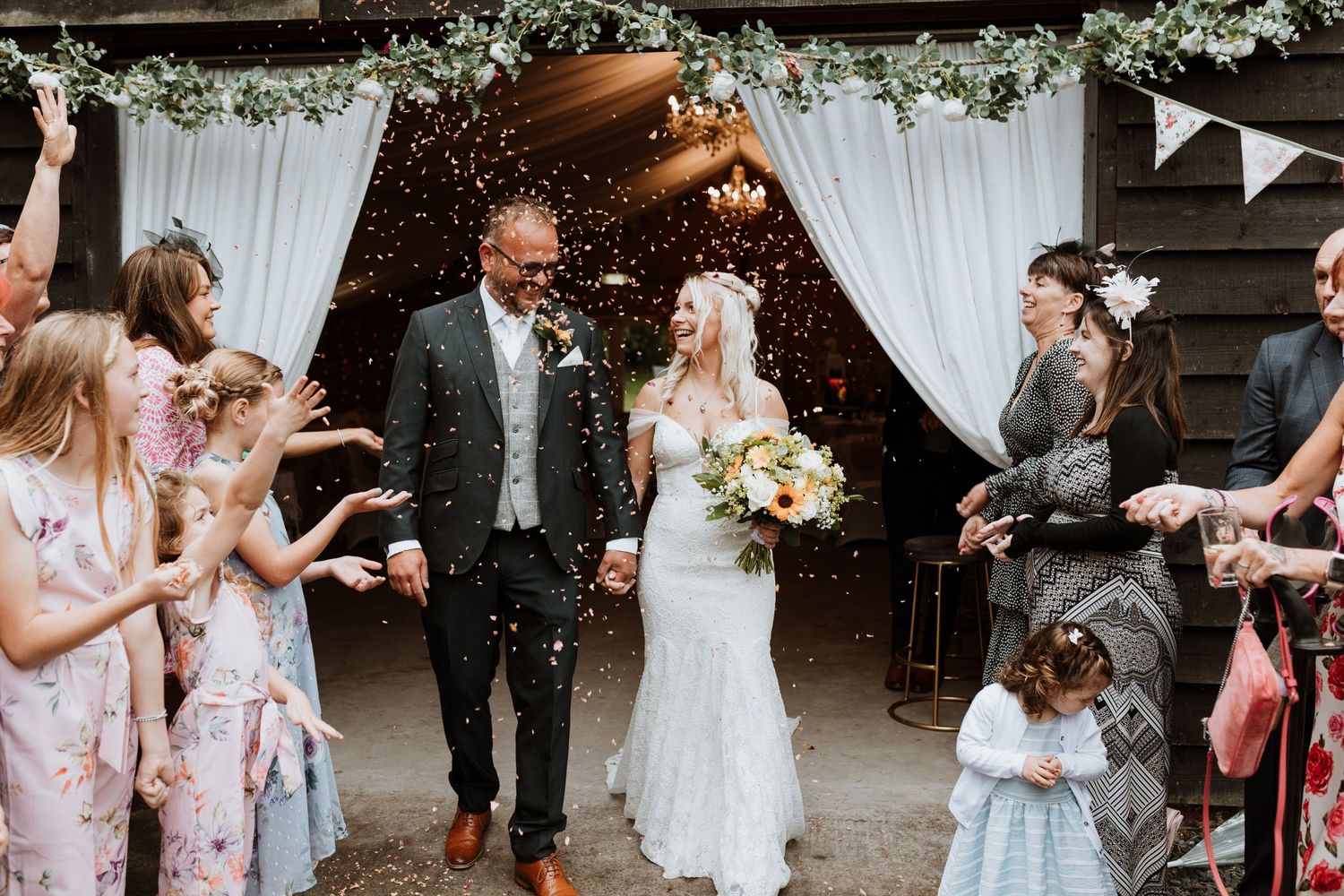 Joyful wedding party throws confetti and celebrates as couple exits rustic barn venue under floral archway at Glyngynwydd Barns.