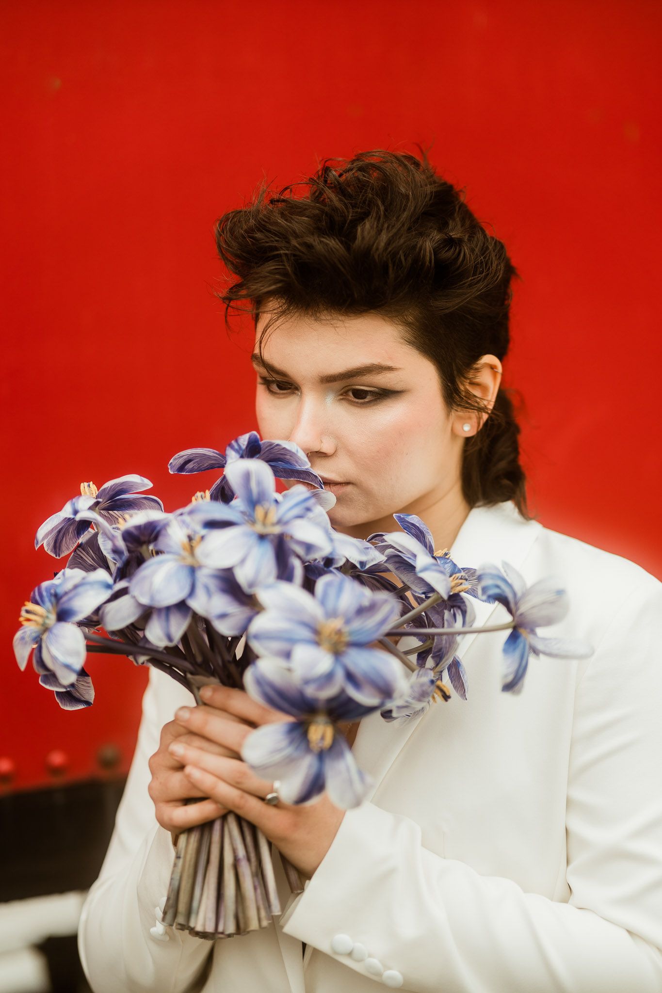 a young woman with short dark hair holding a bouquet on her wedding day against a red backdrop with a white suit on