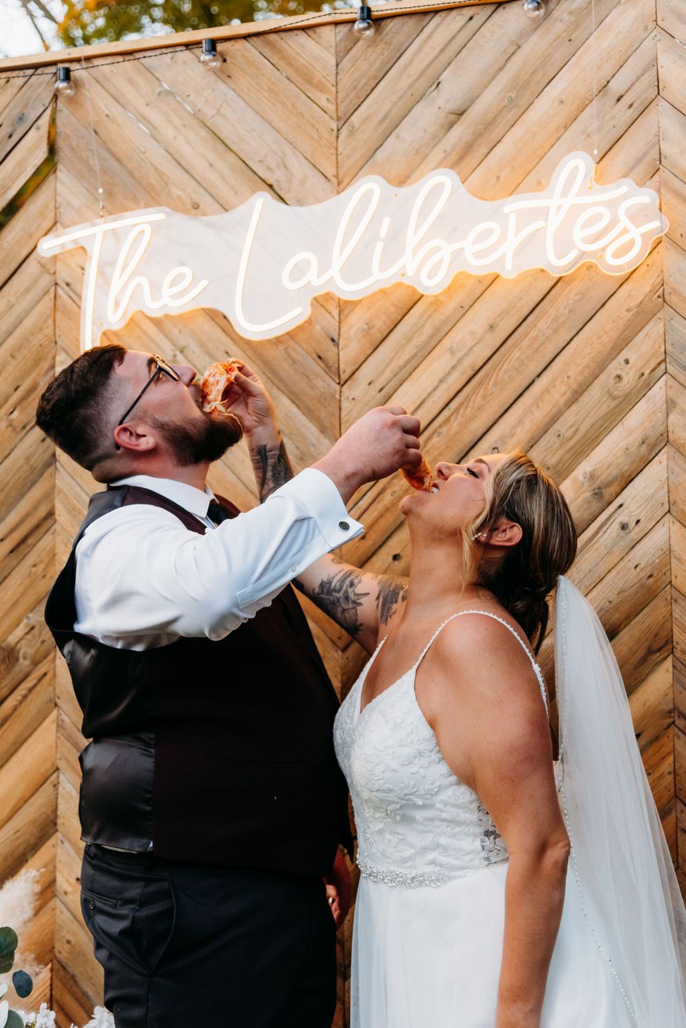 A wedding couple celebrates at their reception with drinks in front of a wooden chevron wall with neon signage.