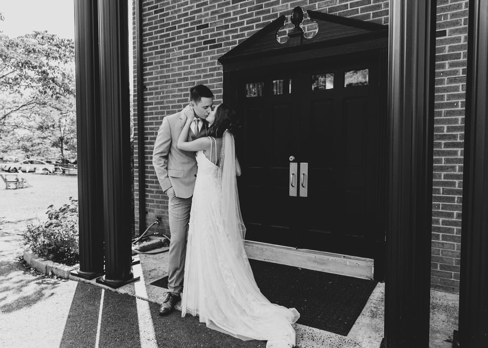 A black and white photo of a couple having a private moment outside a brick church entrance.