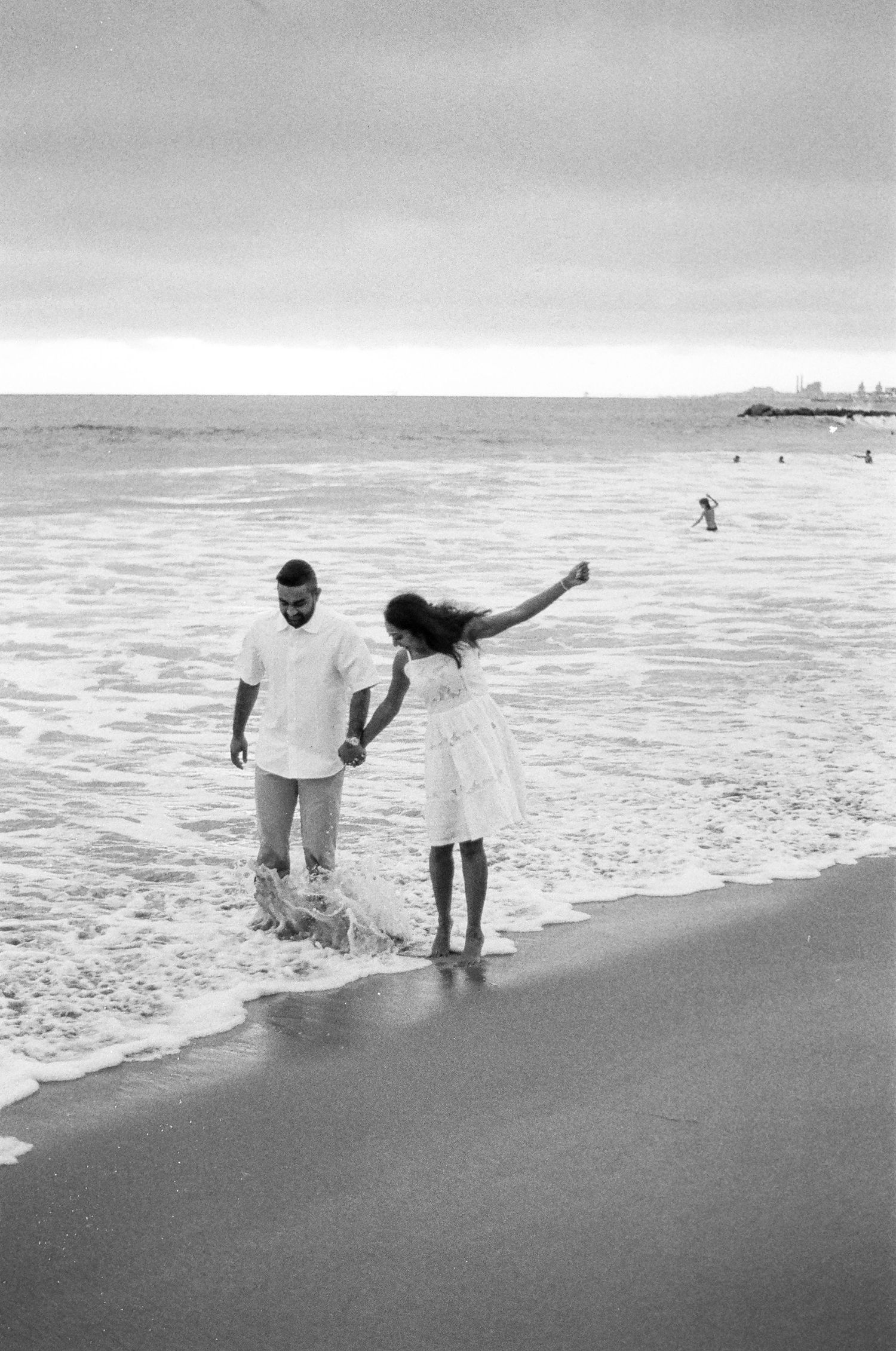 A couple plays together at the beach shoreline in black and white as waves roll in on a cloudy day.