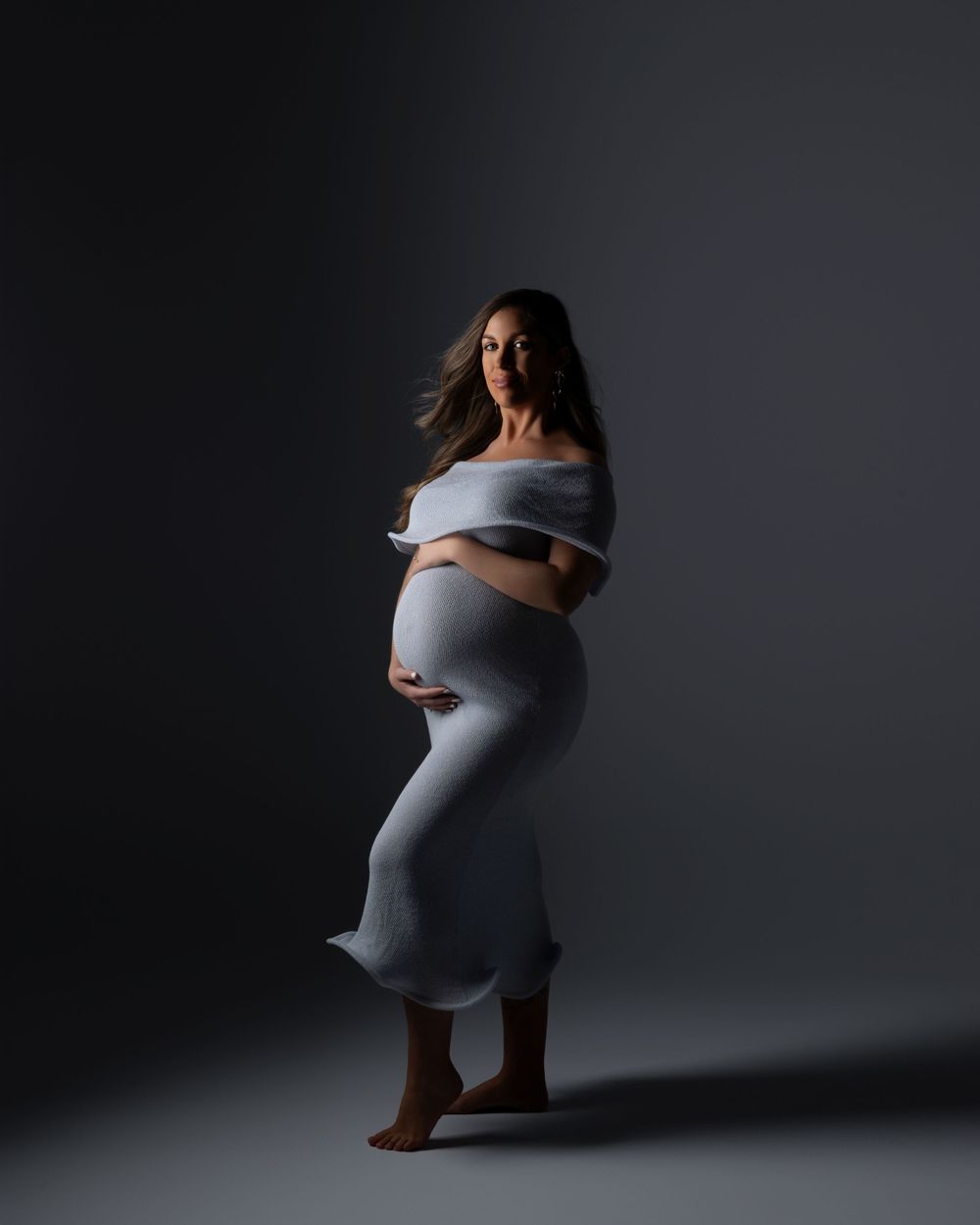 A pregnant figure poses gracefully in a white dress against a dark background in this dramatic maternity portrait.