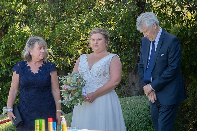 Three people gather outdoors at a wedding ceremony beneath green trees with drinks on a table.
