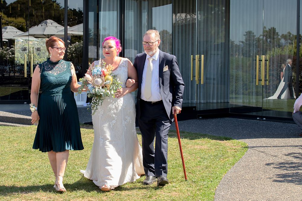 Three people walk together on a path outside a modern building during a wedding celebration.