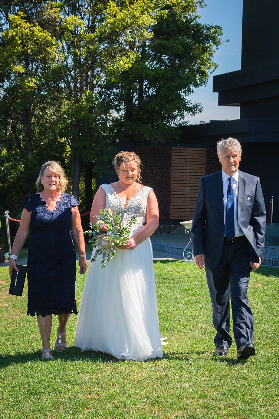 Three people walk across a sunny lawn during an outdoor wedding ceremony.