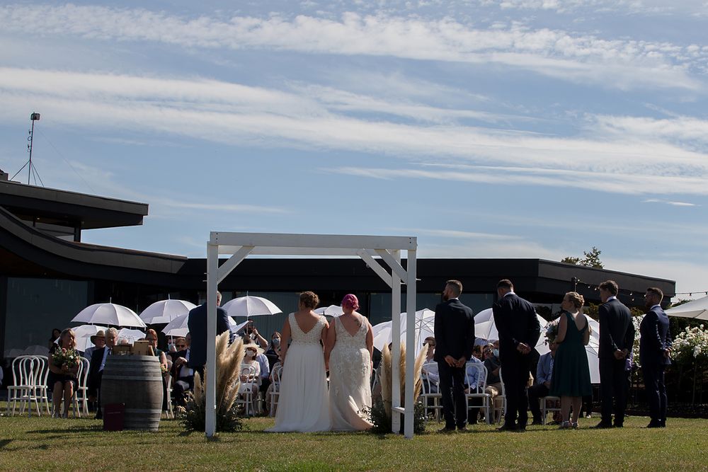 An outdoor wedding ceremony under a white arch with guests holding parasols against a blue sky background.