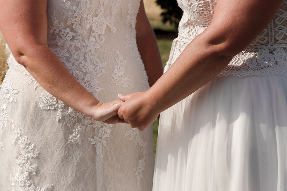 Two people in white lace wedding dresses hold hands behind their backs in an intimate wedding moment.