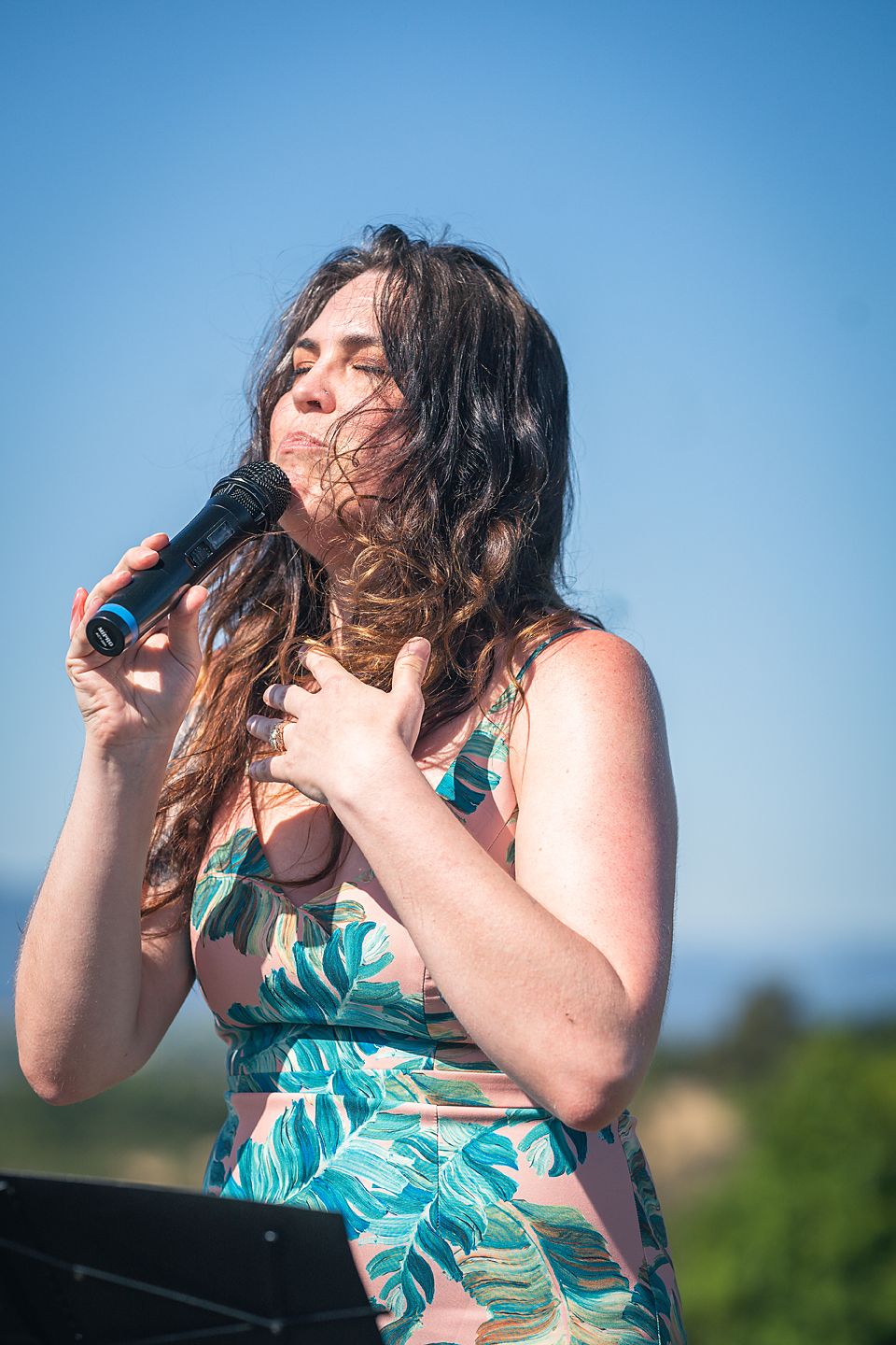 A performer in a green tropical print dress sings passionately on an outdoor stage during a summer performance.