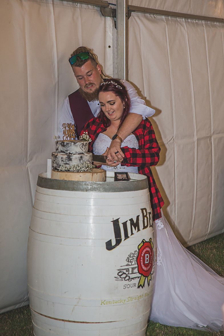 Couple playfully poses with Jim Beam bourbon barrel during wedding photos at an outdoor venue.