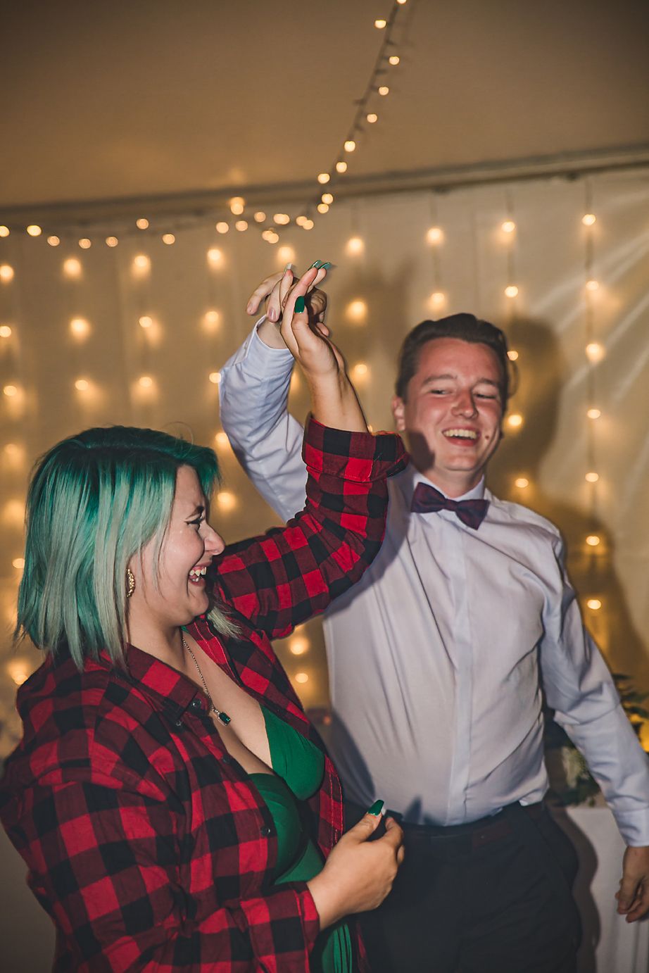 Two people dancing and laughing together under warm string lights at a party, one wearing buffalo plaid and one in formalwear.