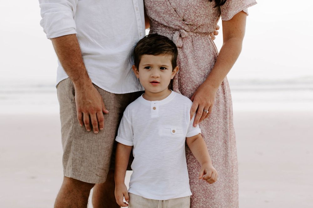Family wearing coordinated white and beige outfits poses together on a bright beach.