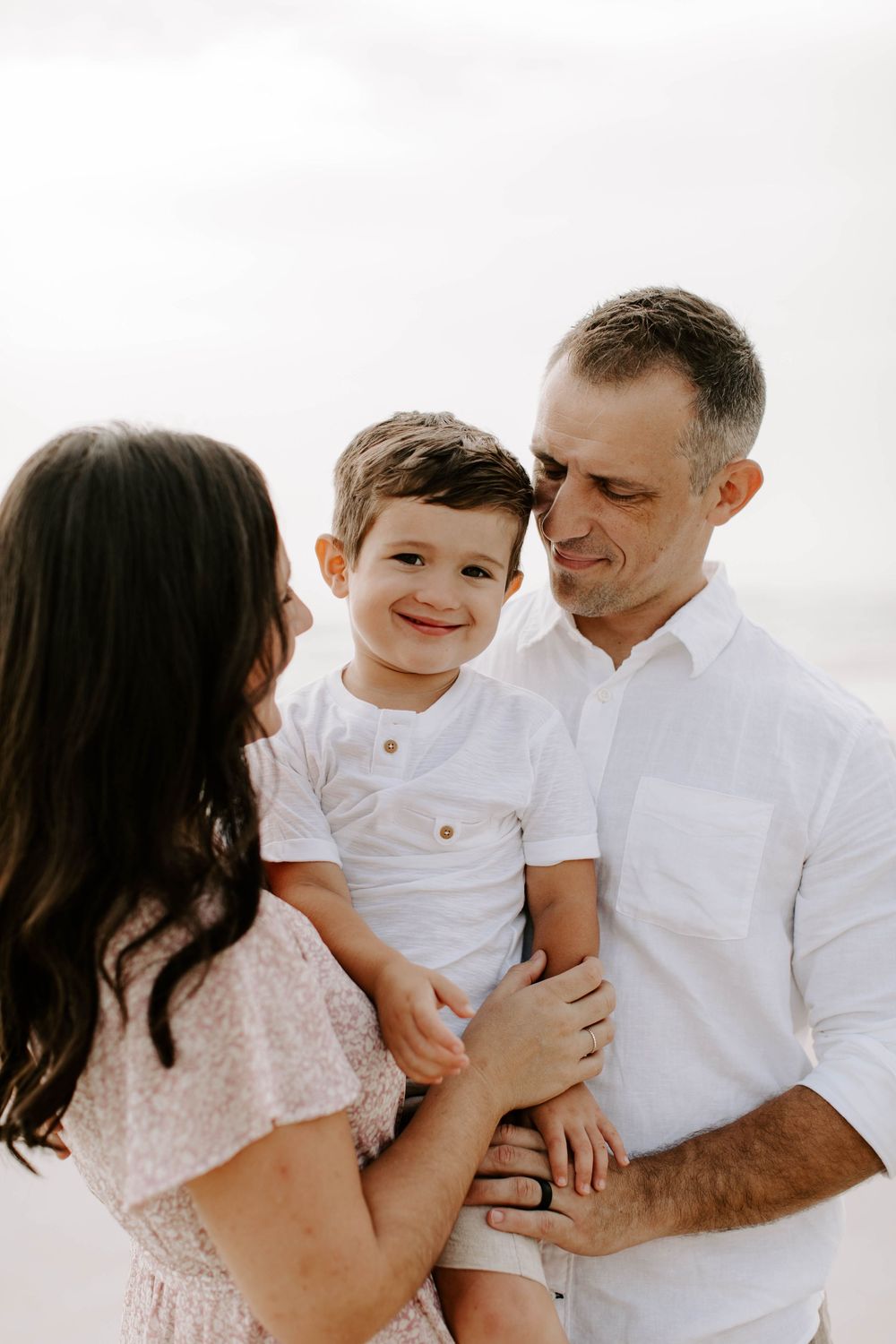 Loving family shares tender moments together during beach photoshoot in white outfits.