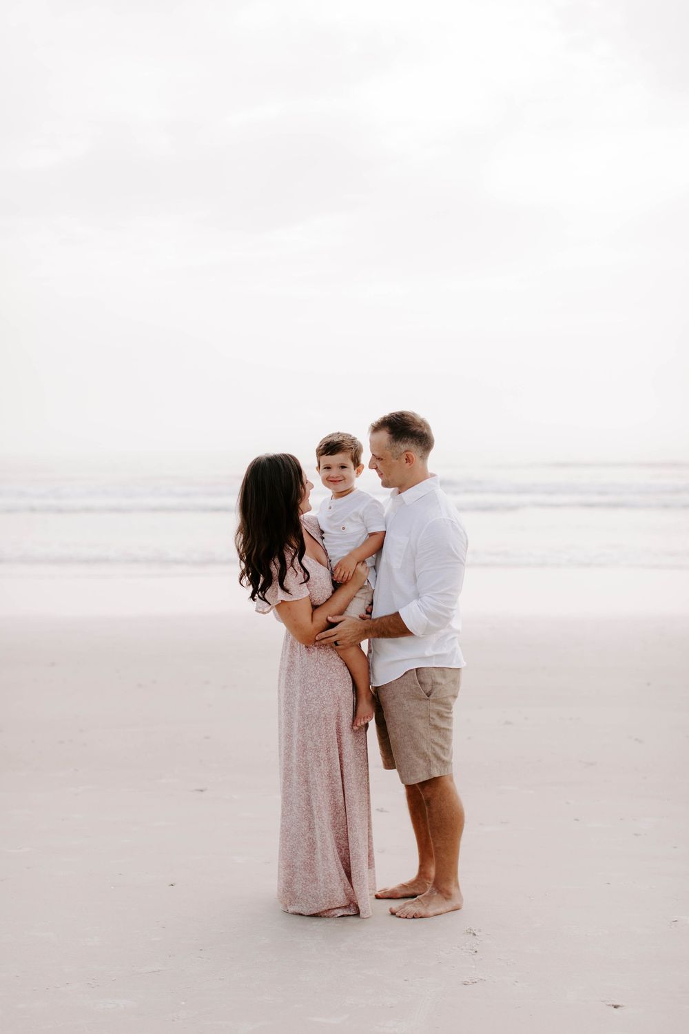 Family dressed in neutral colors stands together on sandy beach during golden hour.