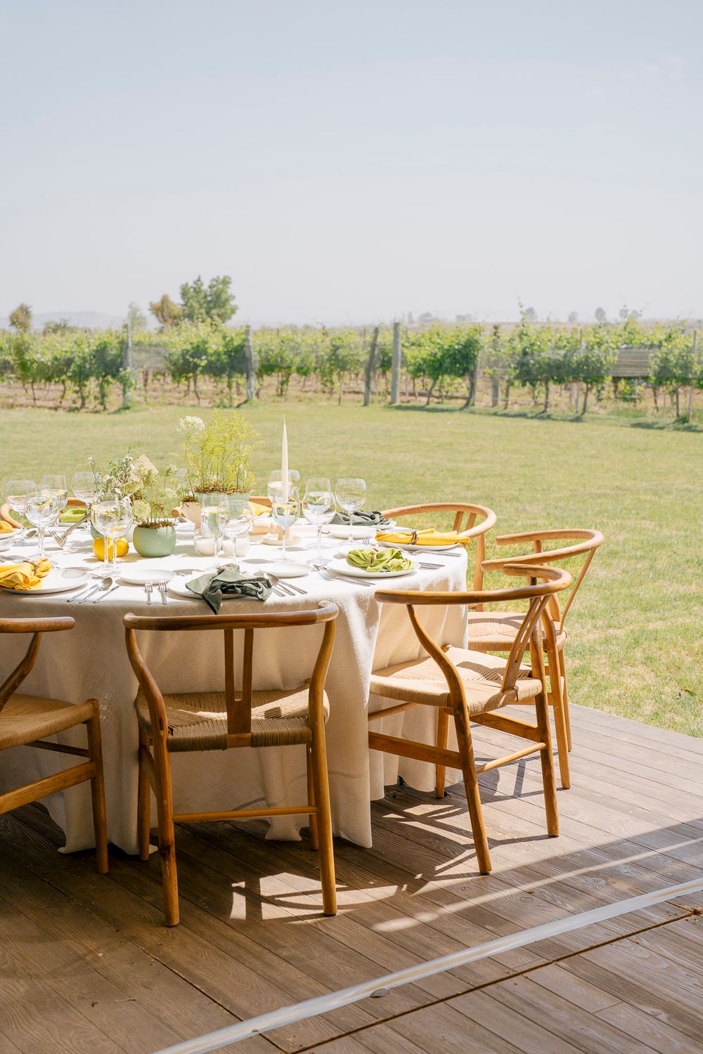 Outdoor dining setup with wooden chairs and round tables on a deck overlooking a sunny vineyard landscape.