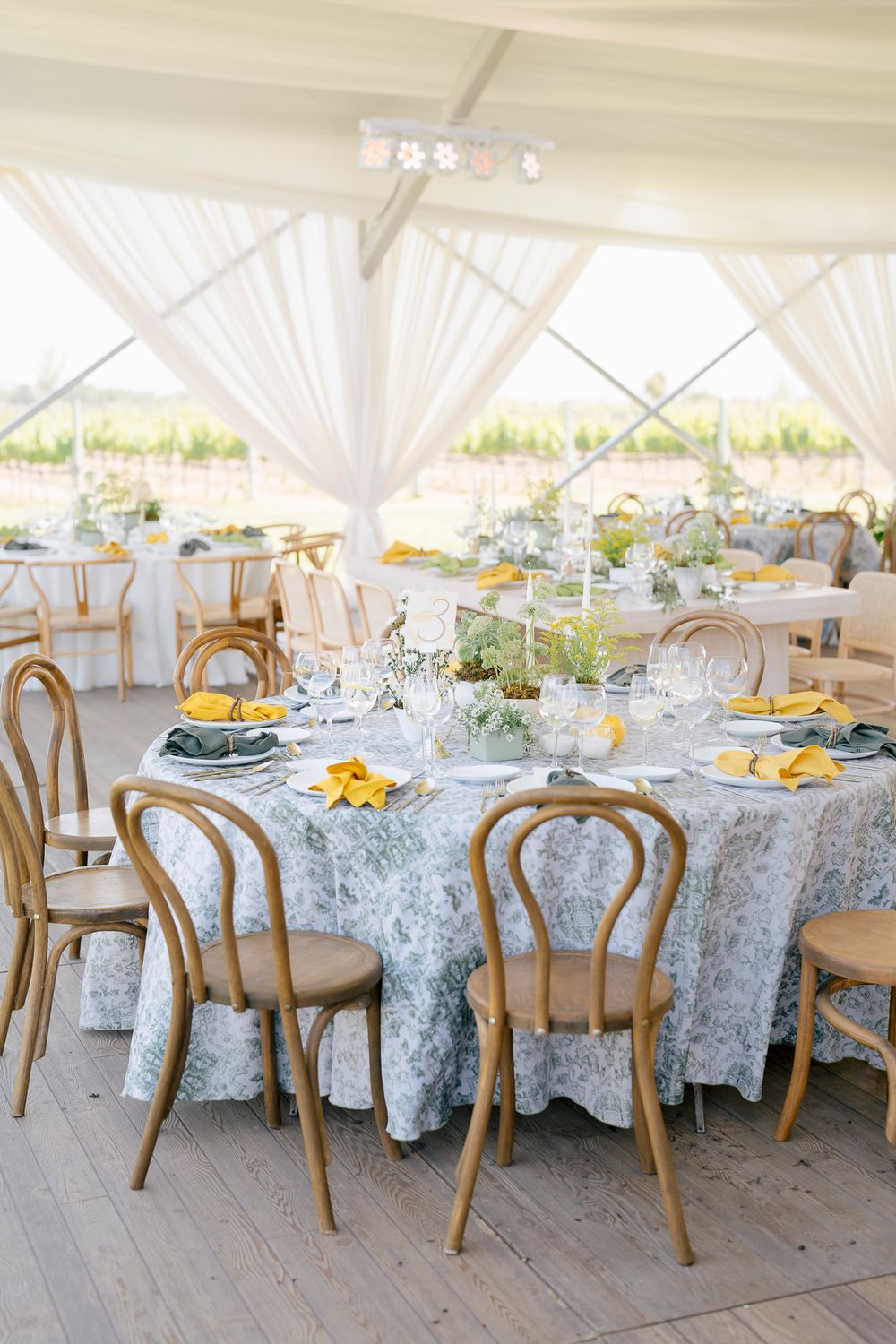 Elegant wedding reception table setup with blue floral tablecloth, wooden bentwood chairs and yellow place settings under white tent.