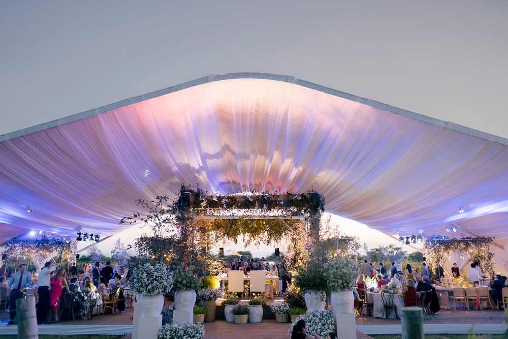 A panoramic view of an elegant indoor wedding reception with white tent ceiling, glowing lights and floral arrangements.