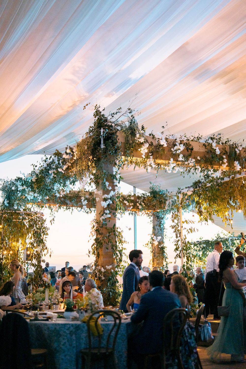 Guests dine under a beautifully decorated outdoor wedding tent with hanging greenery and warm lighting at sunset.