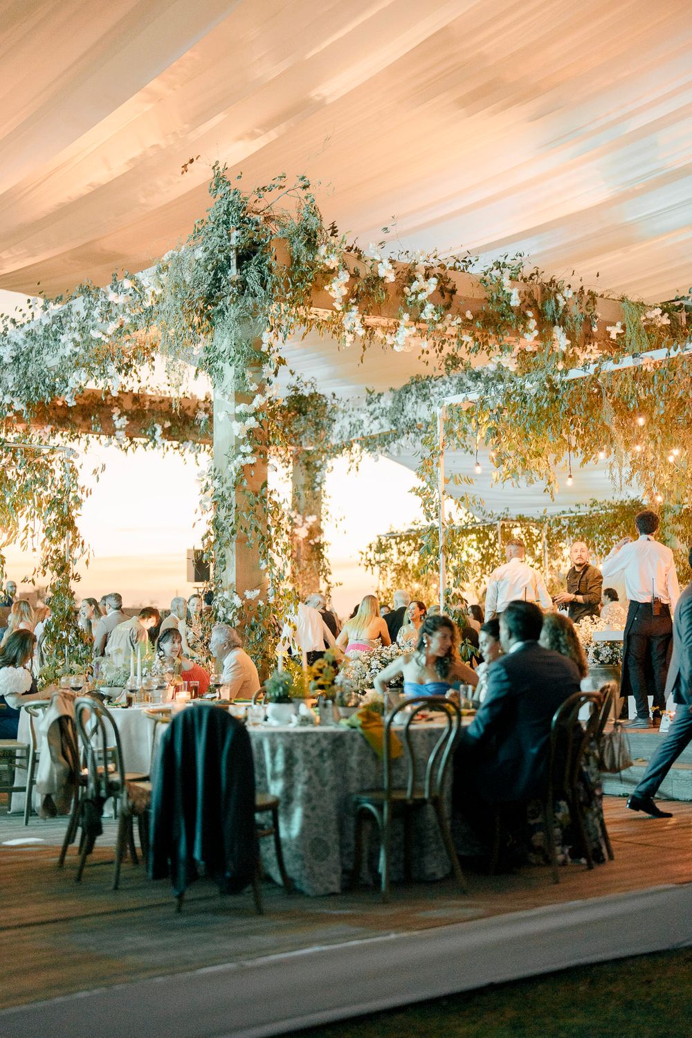 Panoramic view of an elegantly decorated wedding reception tent with floral ceiling arrangements and mingling guests.