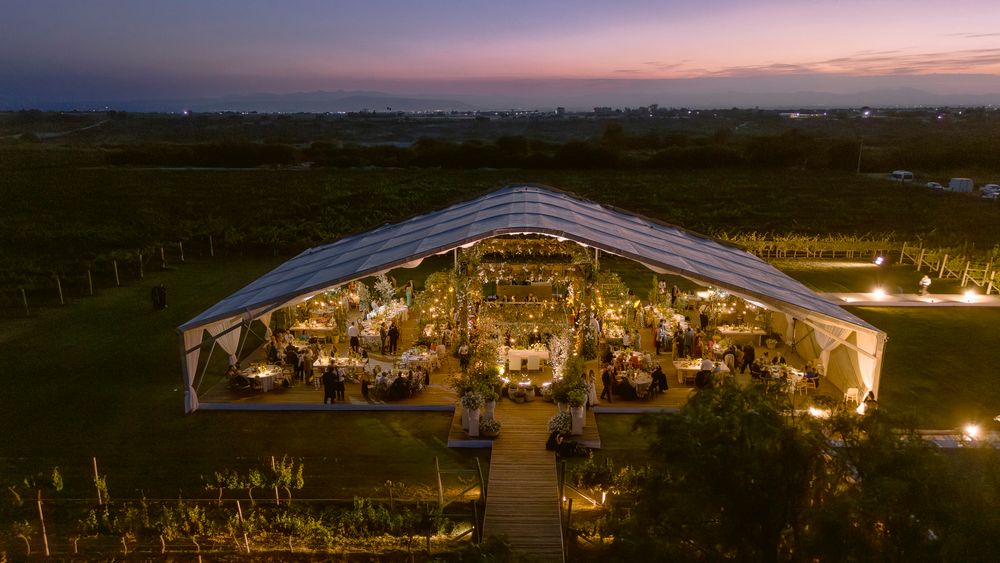 Aerial view of an illuminated outdoor wedding reception tent glowing warmly at dusk in a countryside setting.
