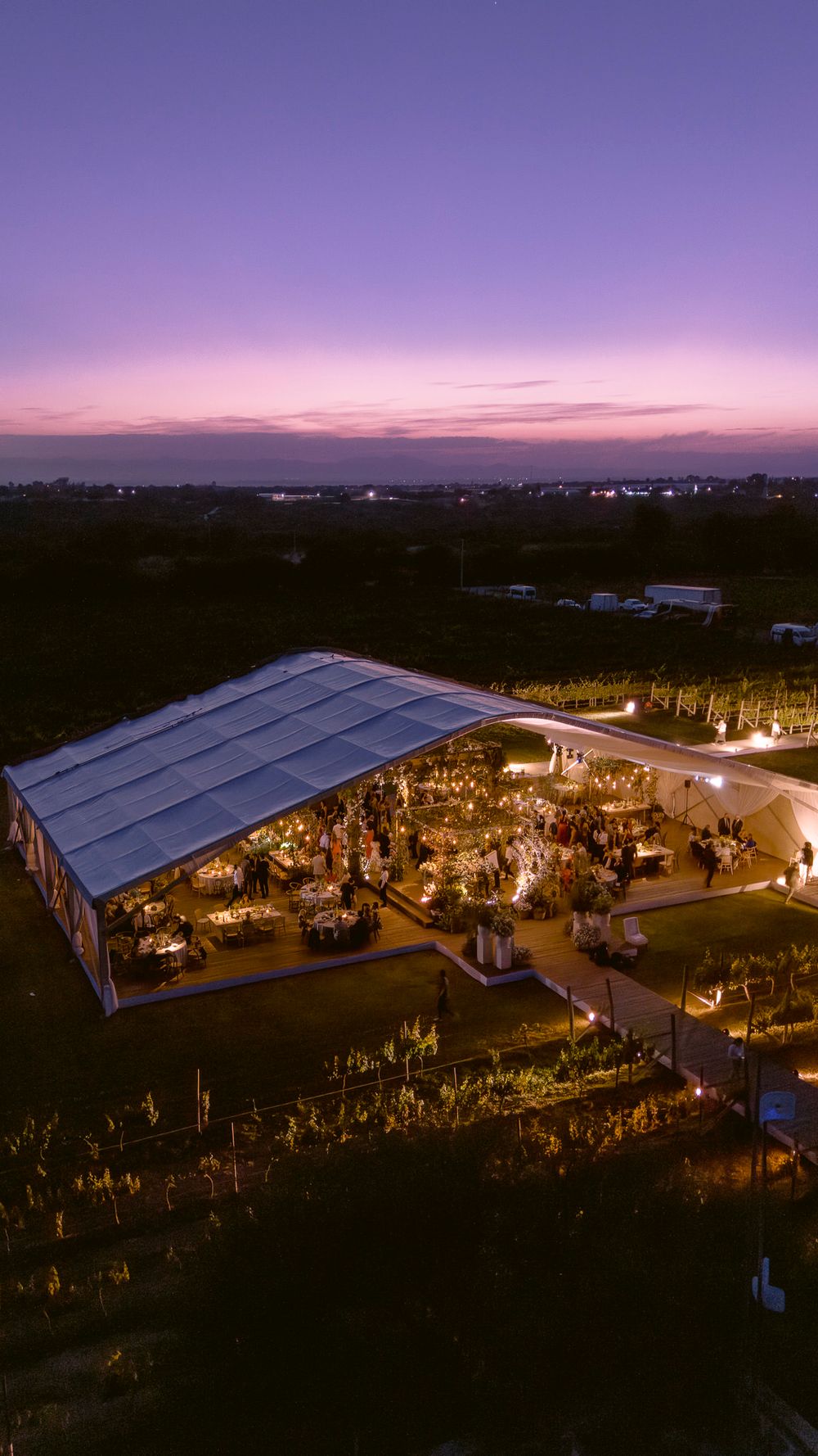 Aerial view of an illuminated outdoor wedding reception tent at dusk with a purple sunset sky in the background.