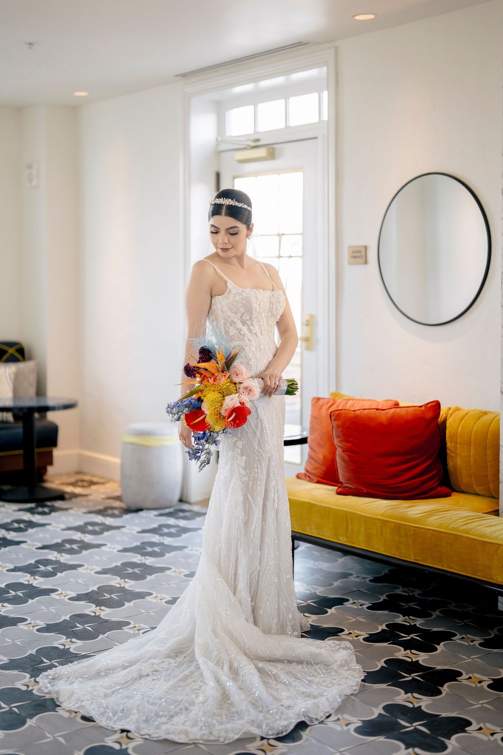 A person in a white lace wedding gown stands on a geometric patterned floor near a vibrant yellow couch.