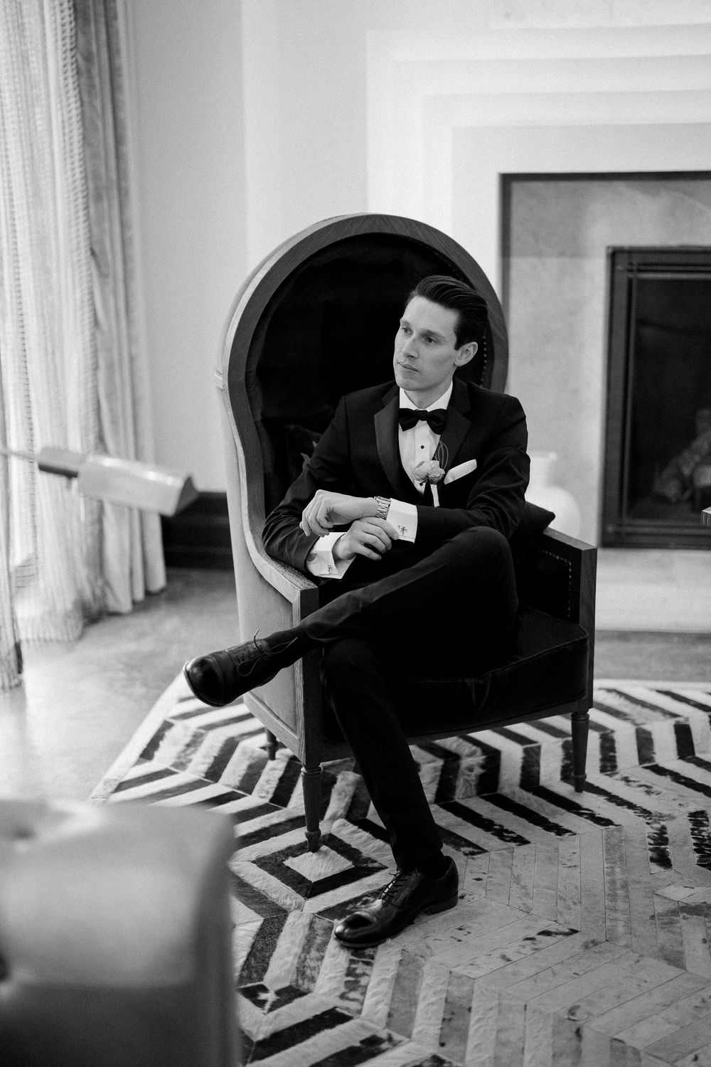 A person in a black tuxedo and bow tie sits elegantly in an armchair against a geometric patterned rug in a formal room.