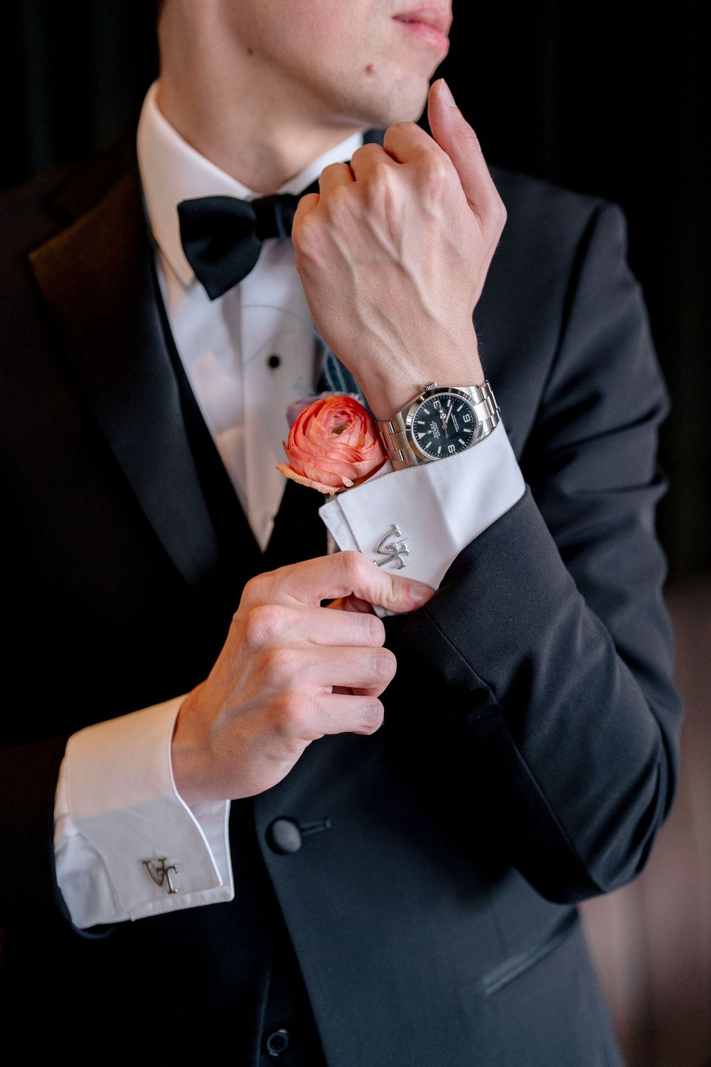Groom in black tuxedo adjusts cufflink while wearing luxury watch and coral boutonniere on lapel.