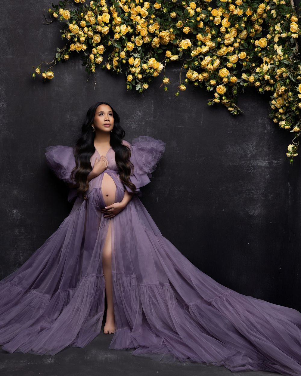 A maternity portrait against a dark wall with yellow flowers overhead, featuring a flowing purple tulle gown.