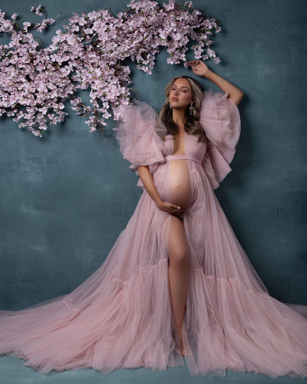 Ethereal maternity portrait with flowing pink tulle gown and cherry blossoms against a moody gray backdrop.