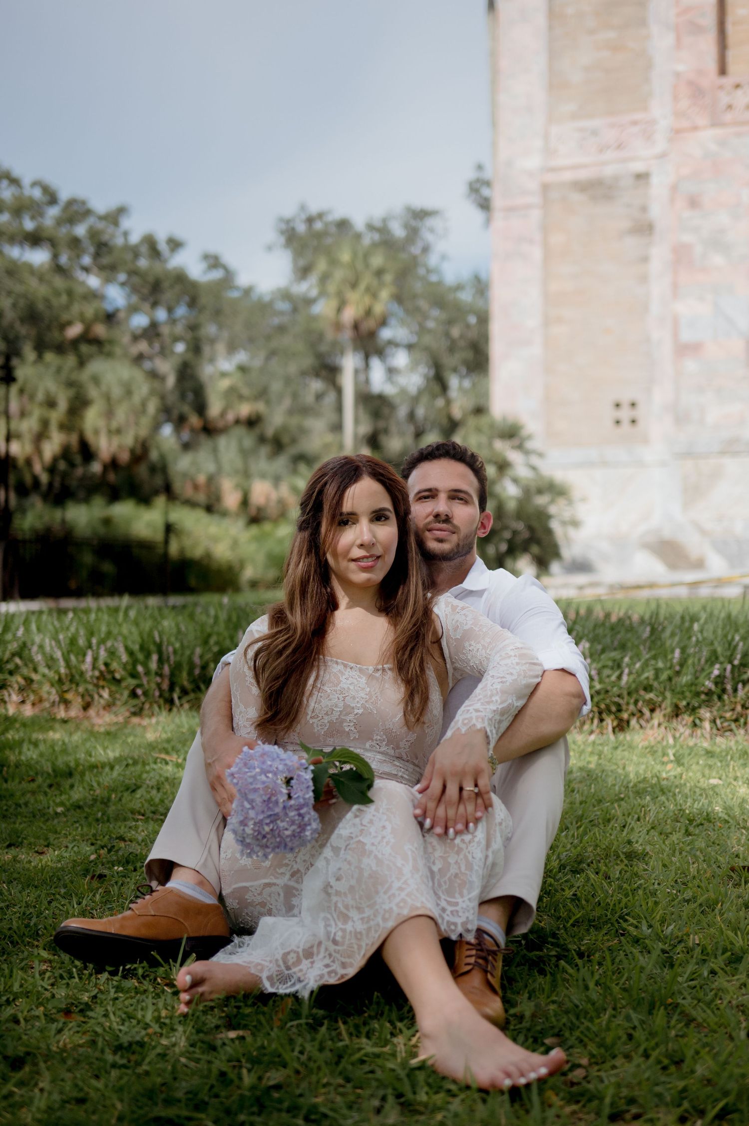A couple relaxes on the grass near a stone building on a sunny day, sharing intimate moments together.