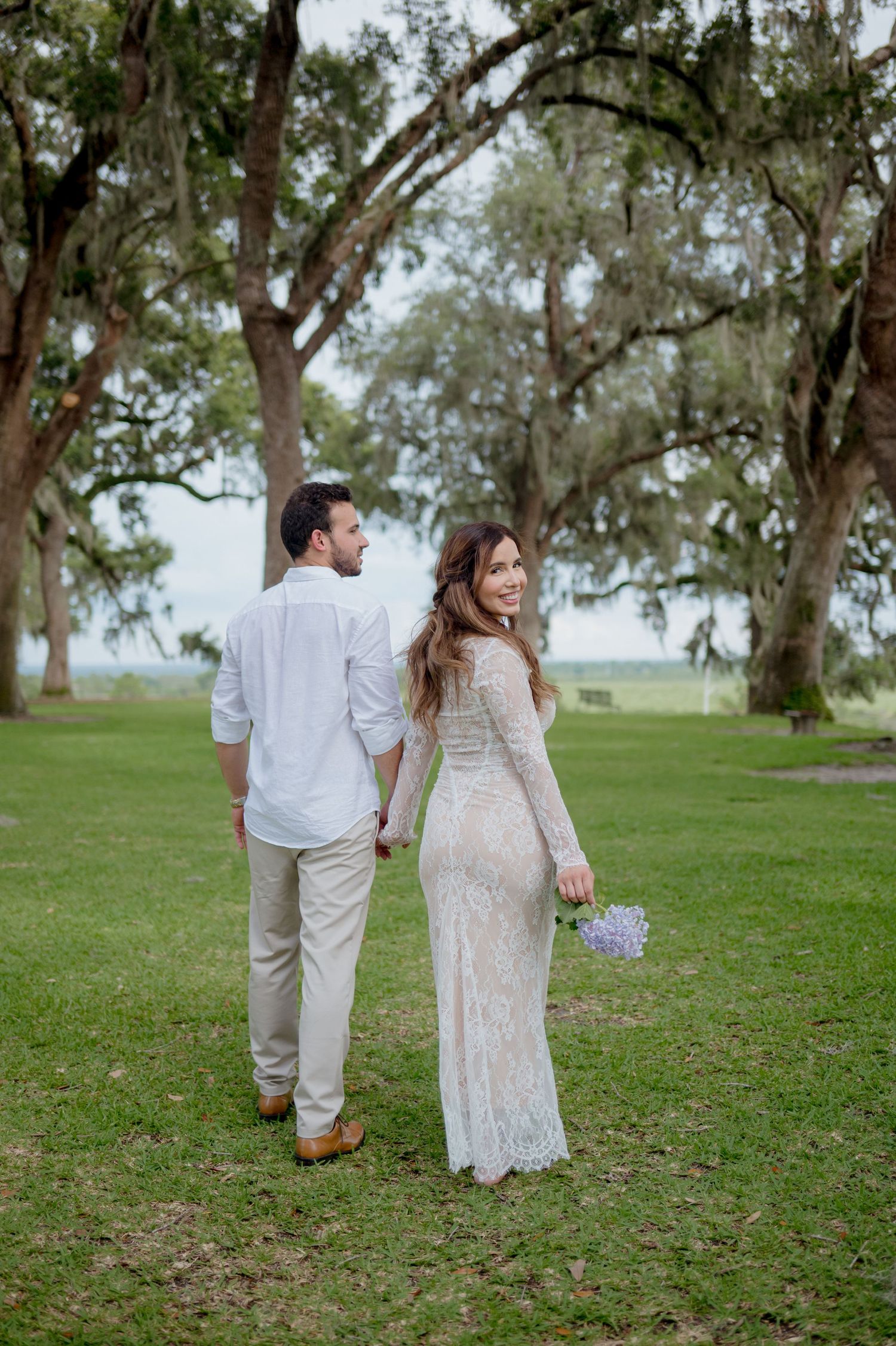 A bride and groom in white attire walk through a scenic outdoor setting with trees in the background.