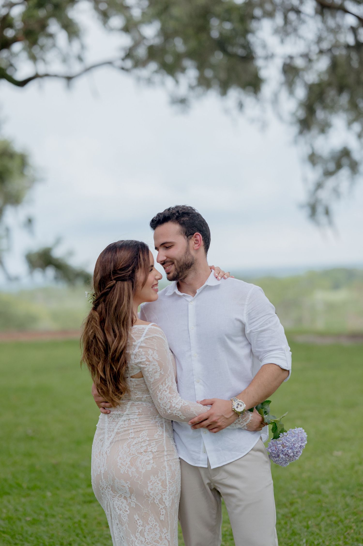 A romantic couple embraces on grassy field during engagement photoshoot with purple wildflowers.