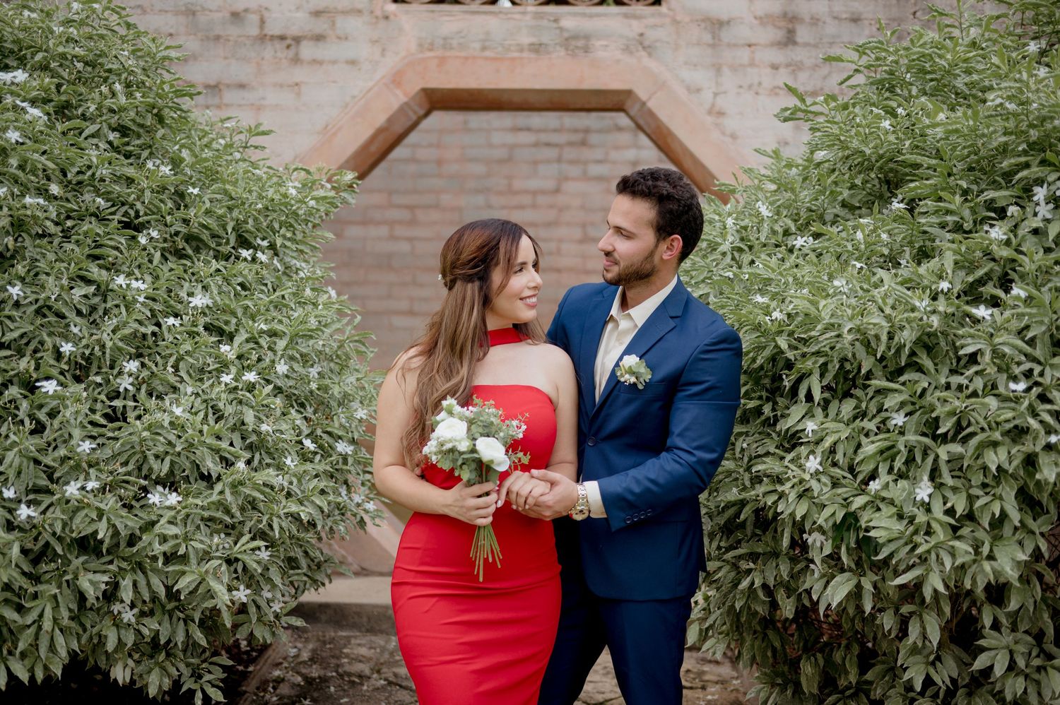 A romantic moment between a couple in a red dress and navy suit amidst lush greenery and a brick wall backdrop.