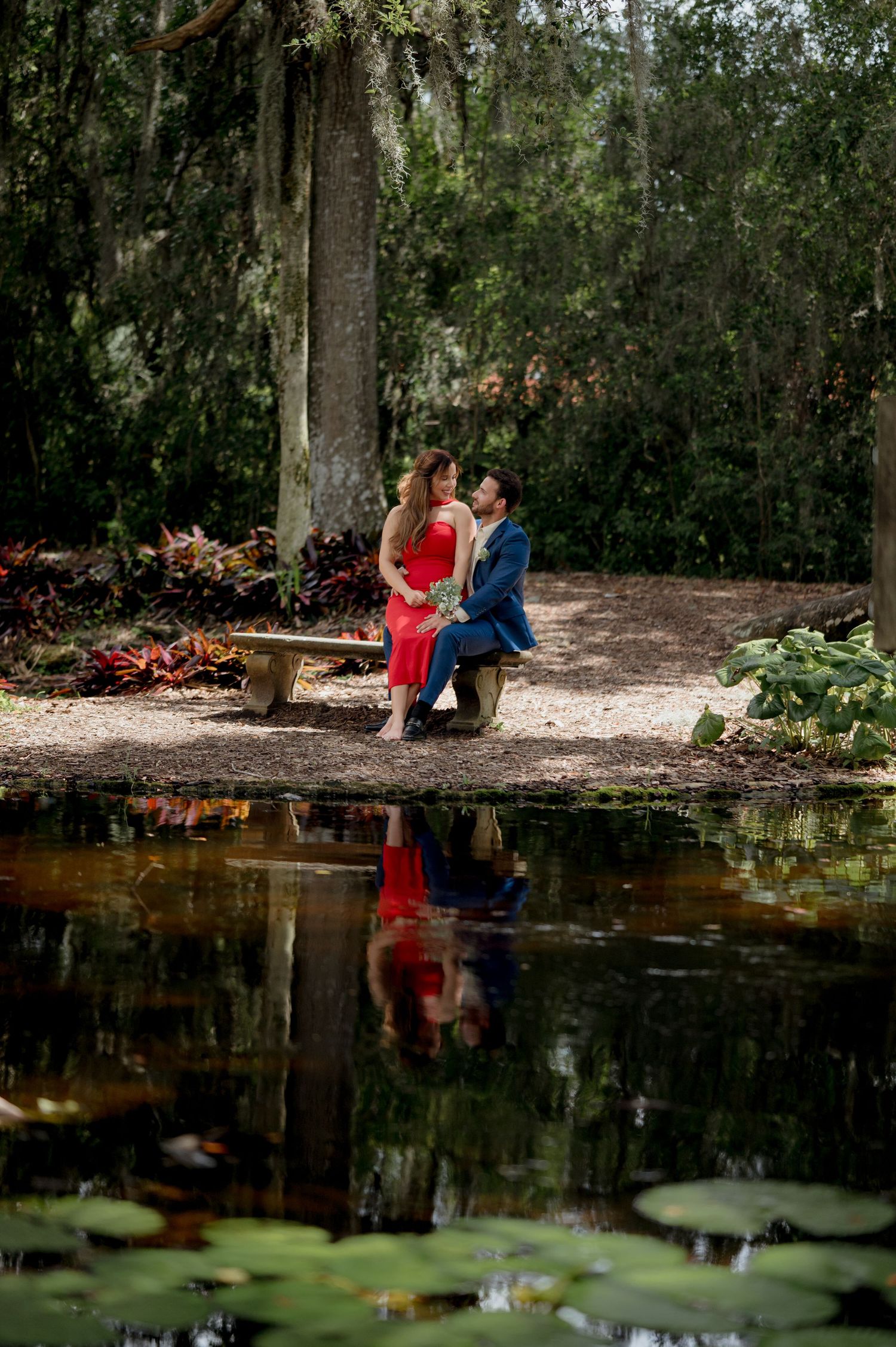 Couple in red and blue outfits sitting on bench by pond with lily pads and Spanish moss in romantic garden setting.
