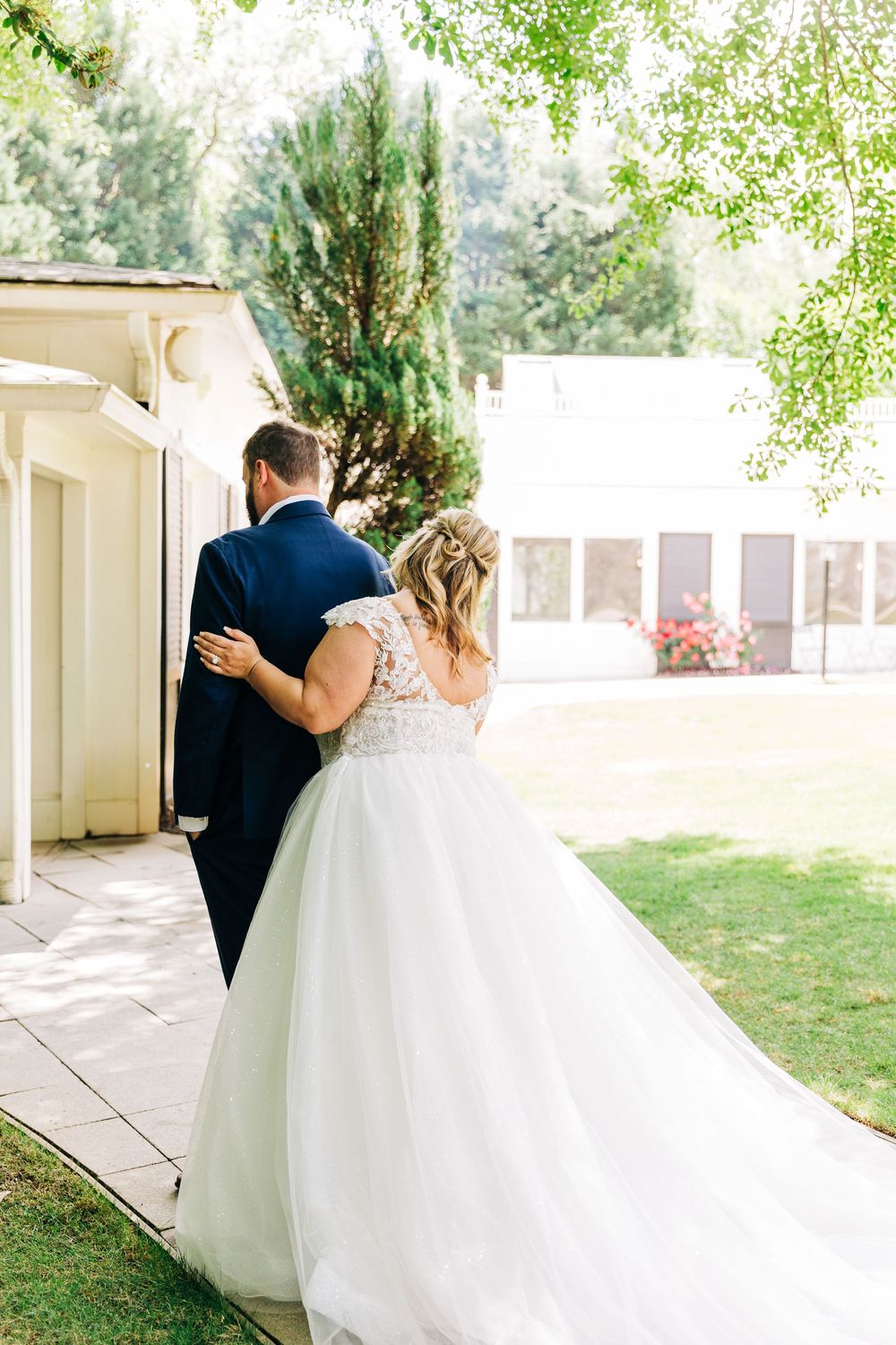 A couple in wedding attire share an intimate moment on a patio walkway surrounded by lush greenery and flowers.