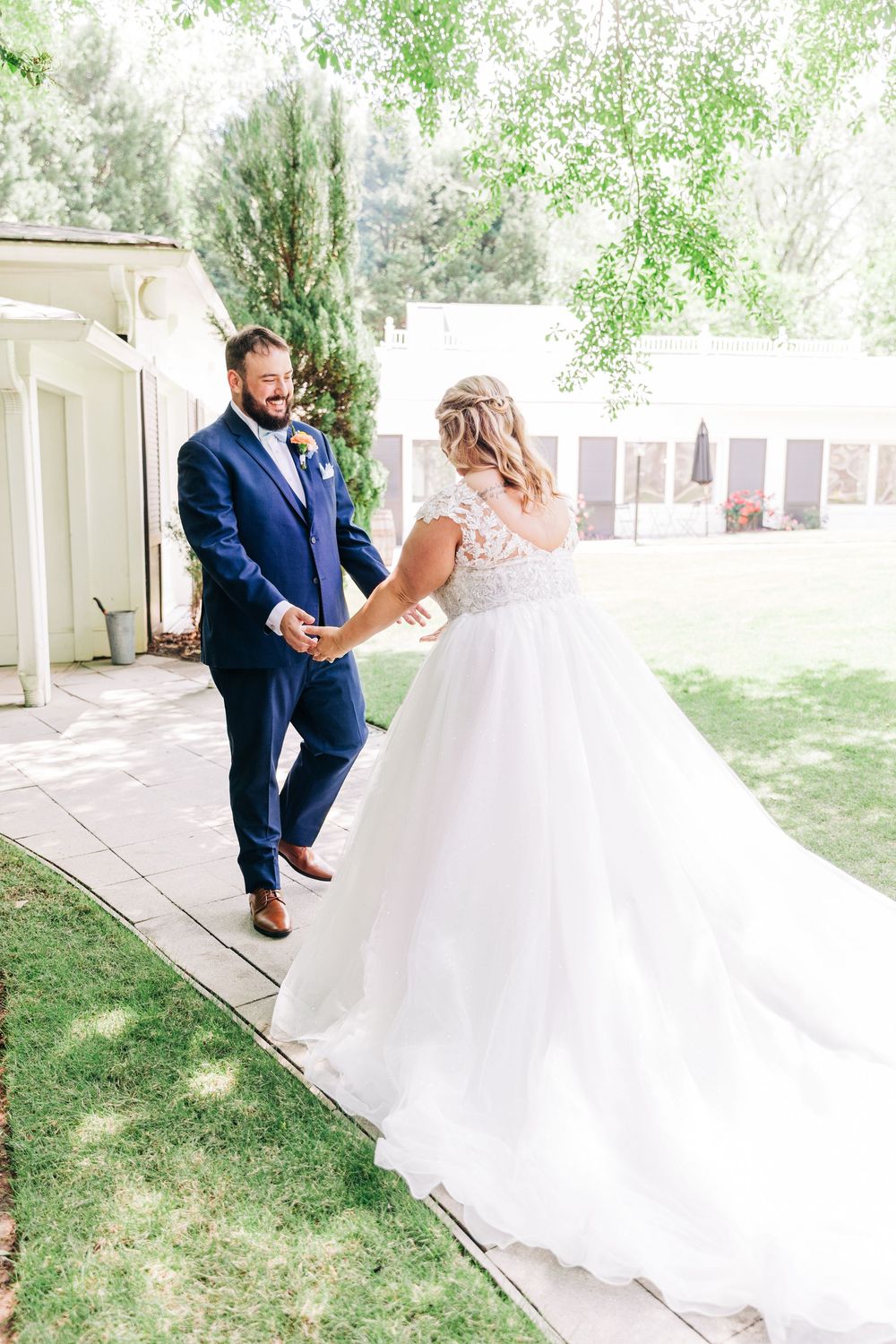 A couple in wedding attire shares a joyful first look moment on a sunlit garden pathway.