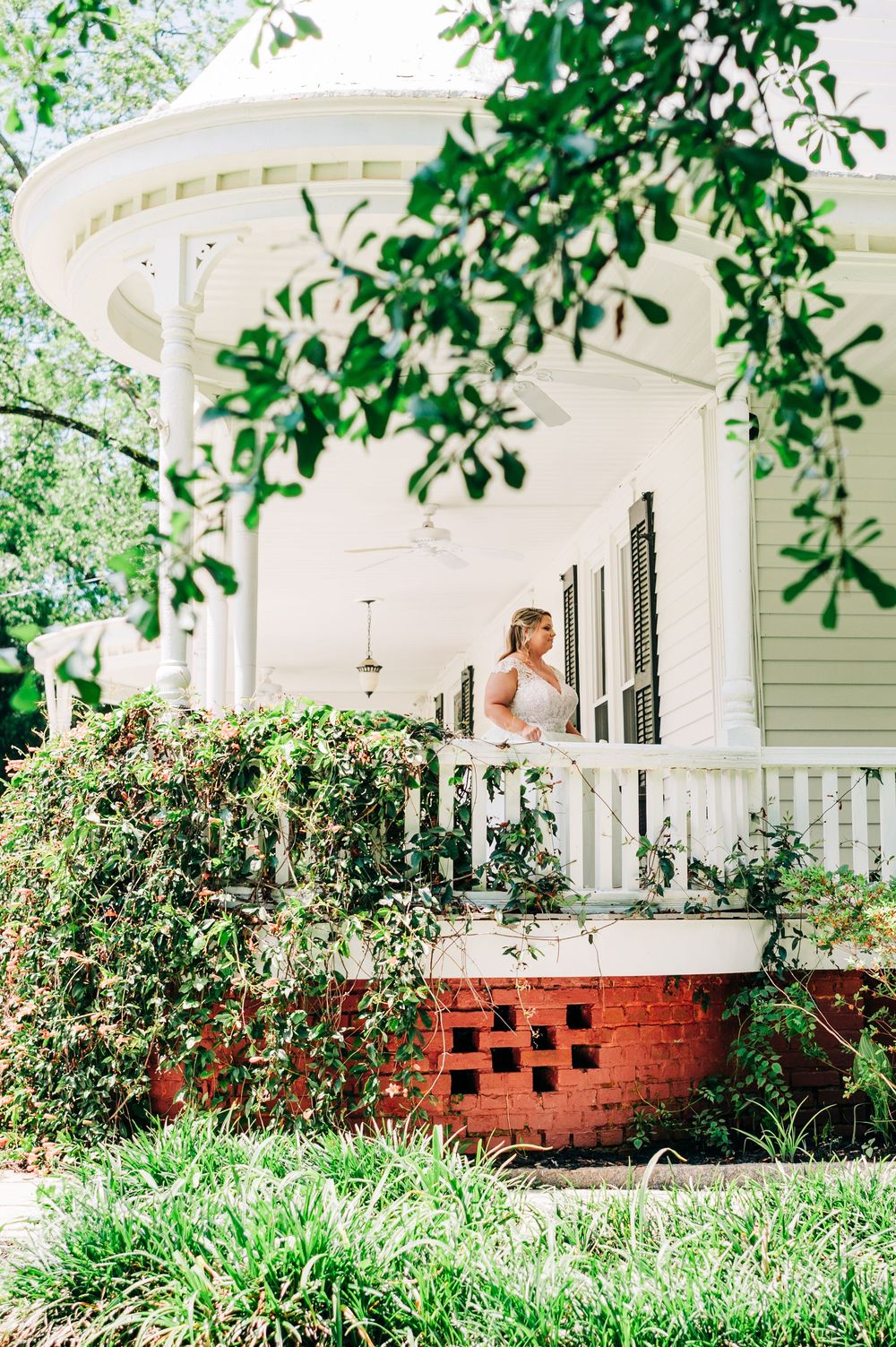 Four sequential images show a white wraparound porch with brick foundation nestled among lush greenery and flowering plants.