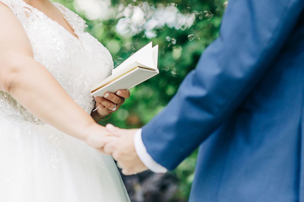 A couple holds hands during a wedding ceremony while reading from a small book outdoors.